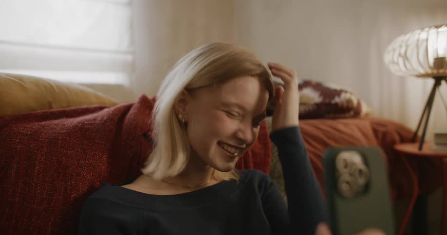 Blonde woman sits by bed making a video call on ihone smartphone