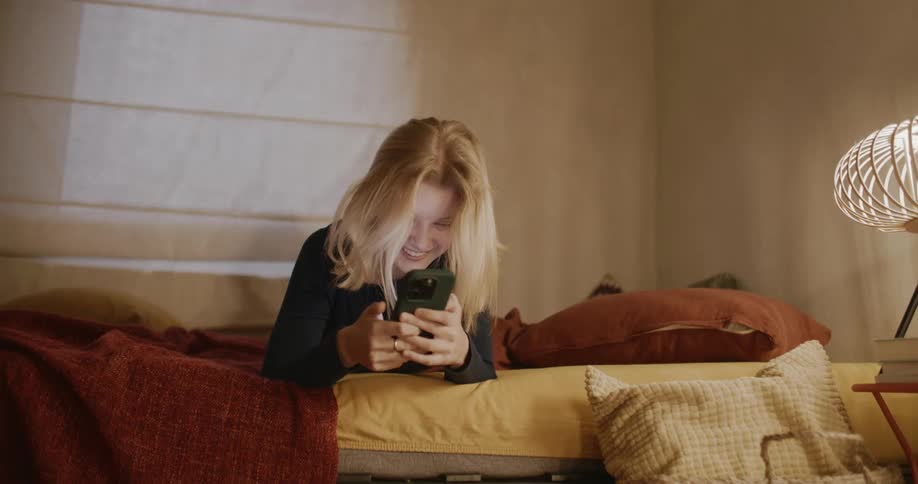 A bedroom scene with a blonde woman laughing while holding a smartphone on the bed
