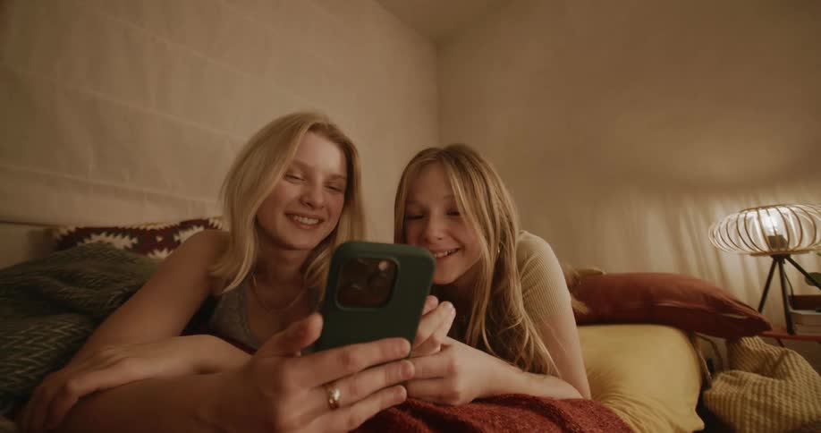 Two teenage friends smile and laugh at a phone in the bedroom