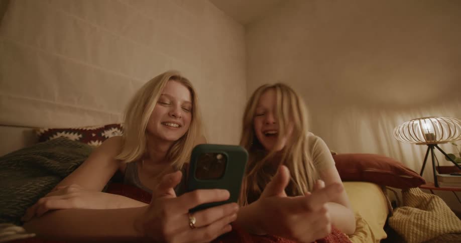 Two girls lying on a bed talking and laughing while looking at a smart phone
