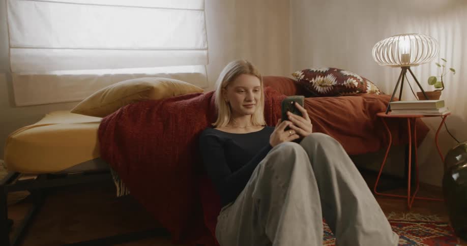 Young woman sitting on bedroom floor laughs while viewing smartphone screen