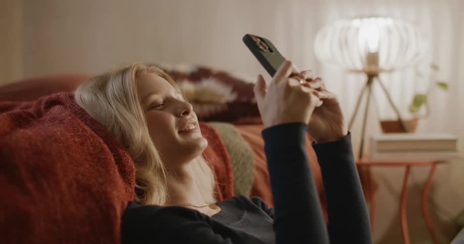 Portrait of a blonde woman sitting on the floor leaning against the bed smiling and using her cell phone