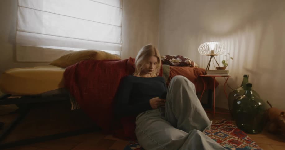 A woman sits by her bed using a cell phone