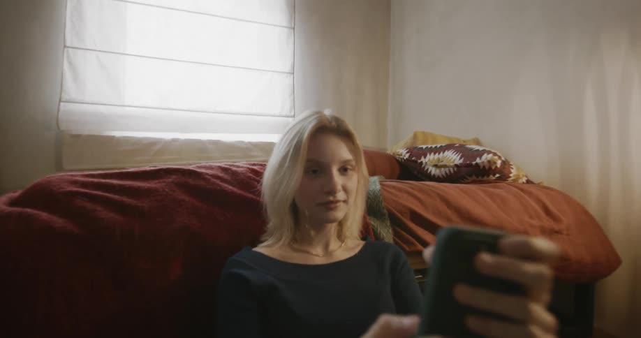 A blonde lady using a mobile phone while seated on the floor near the bed during the day
