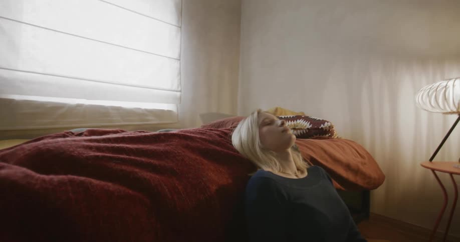 A woman sitting on the floor next to a bed with a red blanket in a softly lit room with a window