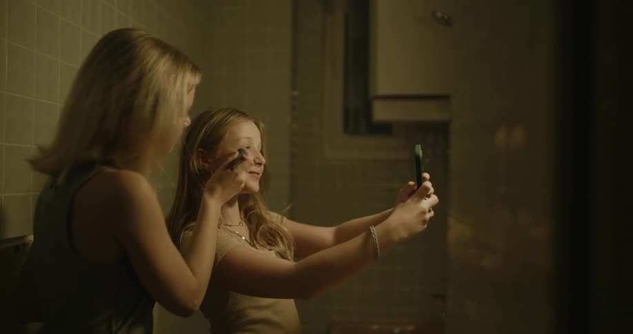 Teenage girl smiling and taking selfies inside the restroom while another girl retouches her face with a brush