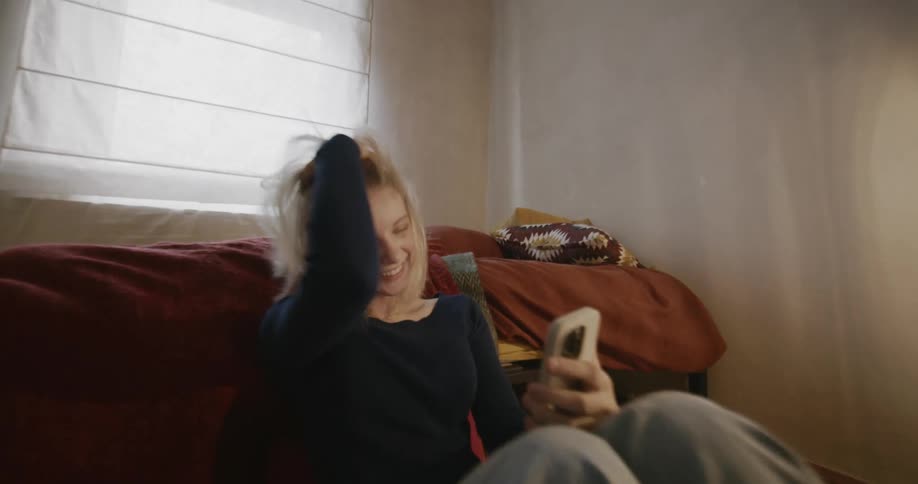 Young blonde woman sitting next to the bed using her cell phone