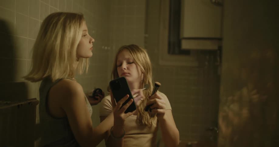 Girl holds a smart phone while another girl retouches her makeup inside the restroom
