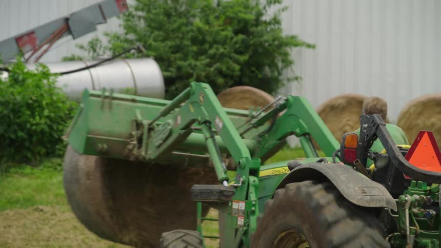 A person operating a green tractor lifting a round hay bale in front of a white barn
