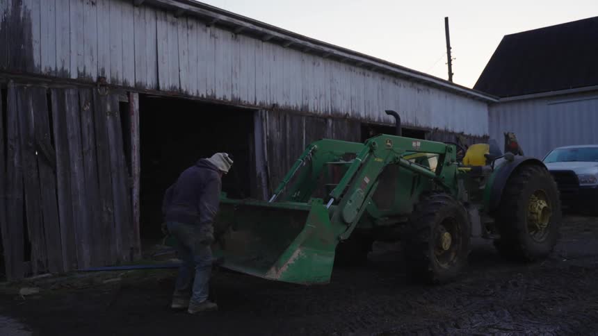 A person working near a green tractor with a front loader parked near an old wooden at morning