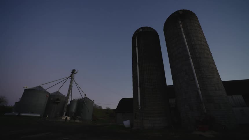 A farm scene in the evening with two silos and several grain storage units