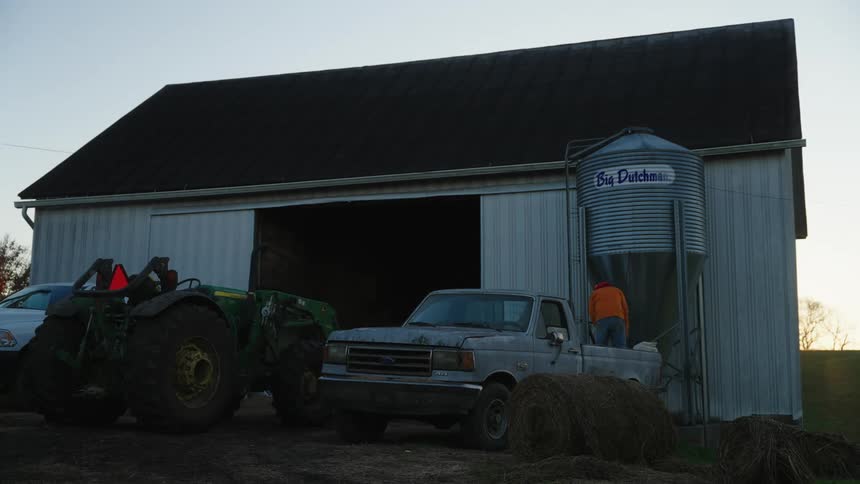 A barn with open doors with a green tractor and a blue truck during dusk