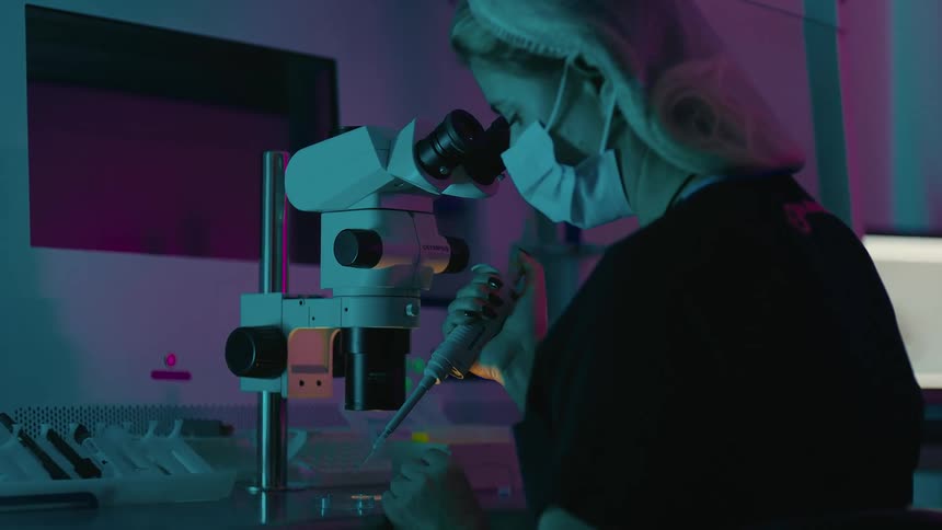 A person in a laboratory with a pipette and a microscope working with liquid samples in purple lighting