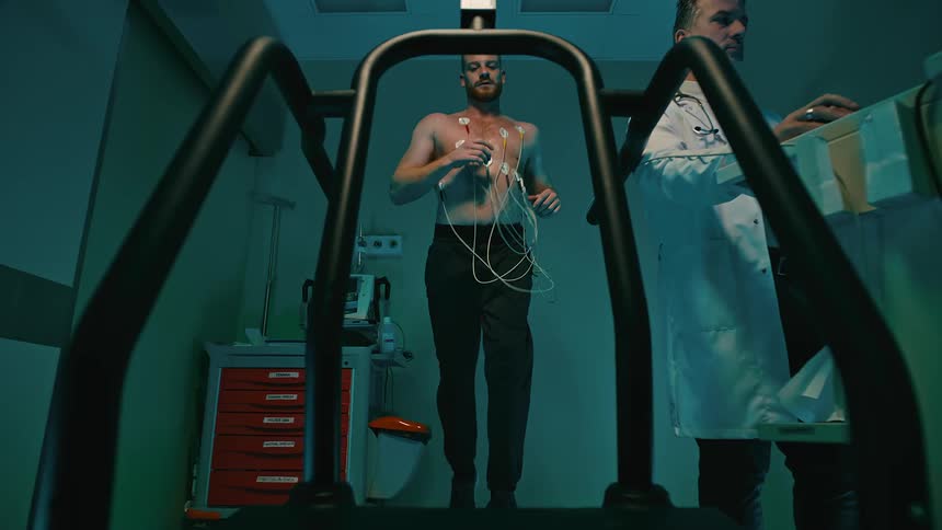 A person is running on a treadmill with multiple electrodes attached to their chest while a doctor observes