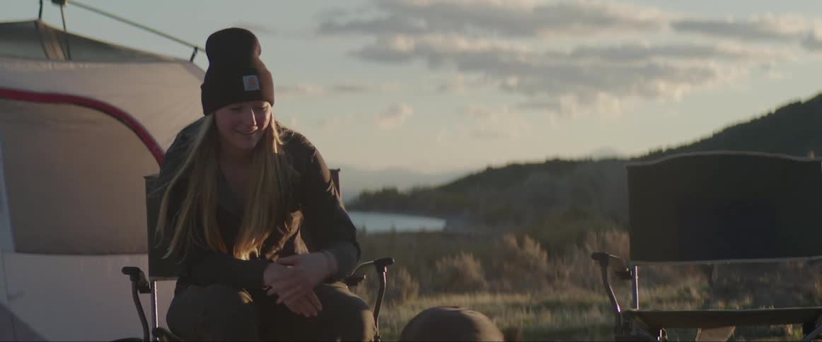 A woman wearing a beanie sitting on a folding chair in a grass field in front of a tent pets a dog