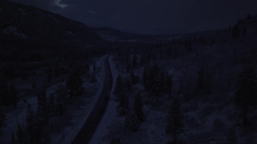 Two rugged vehicles driving single file down a highway surrounded by snow dusted wilderness trees in a mountain valley after sunset just before dark