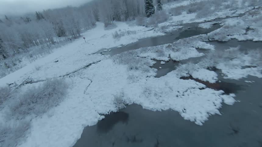 Flying perspective between frozen snow covered pine trees along a stream and over a frozen lake in a valley surrounded by cloud covered mountains