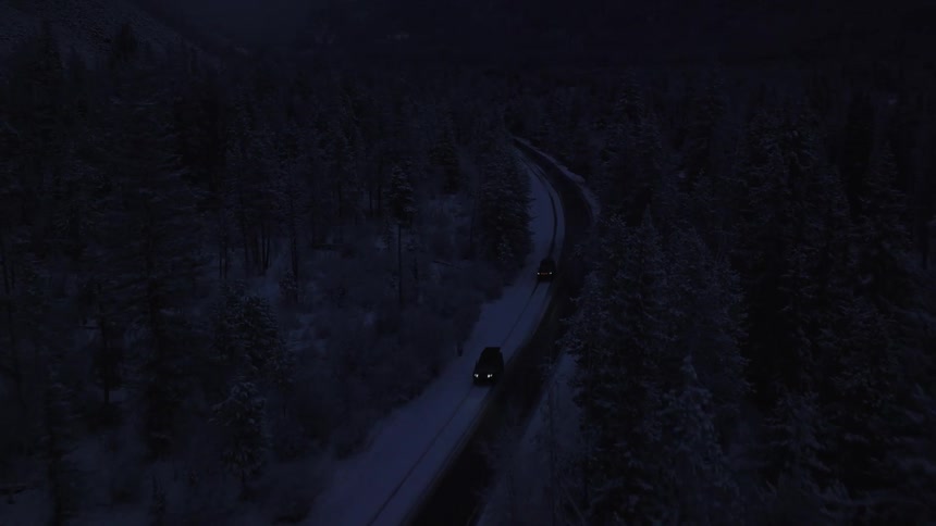 Two cars driving on a rural street surrounded by pine forests and there is snow on the ground