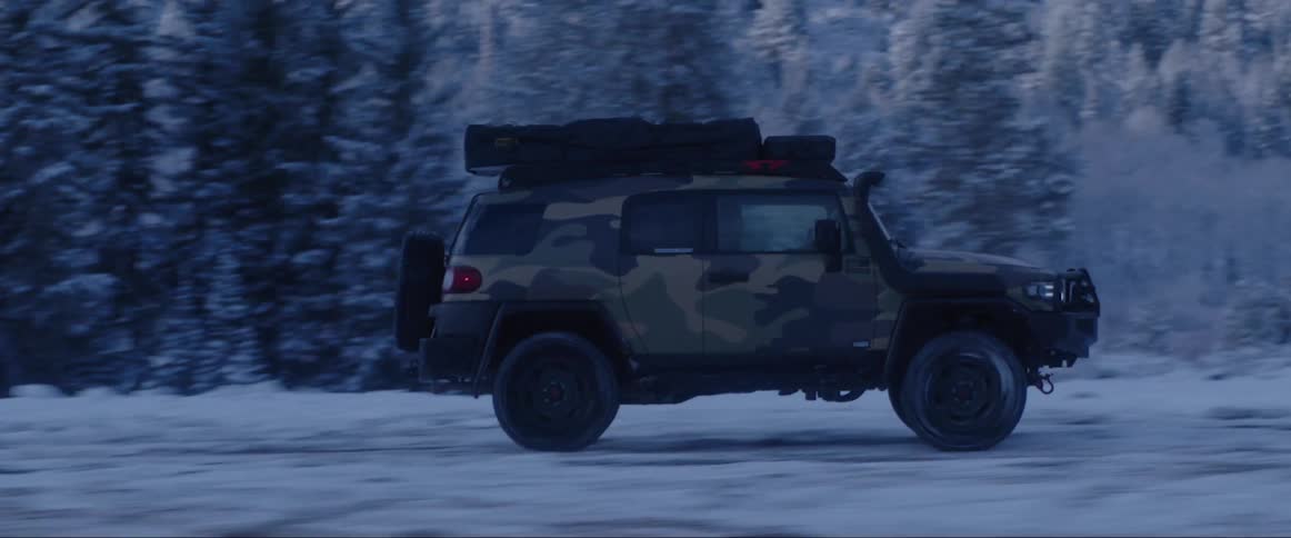 A camouflaged suv drives on a snowy terrain with a forest in the background