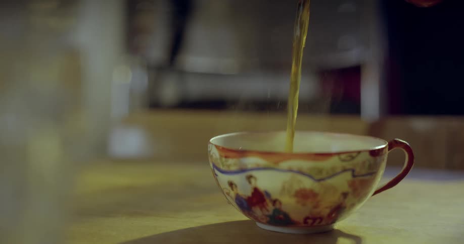 A persons hand picking up a tea pot and pouring tea into a an ornate painted china cup