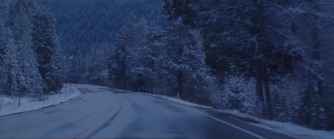 Street level driving view along road through a snow covered forest of conifer trees surrounded by snow covered tree covered hills after dusk
