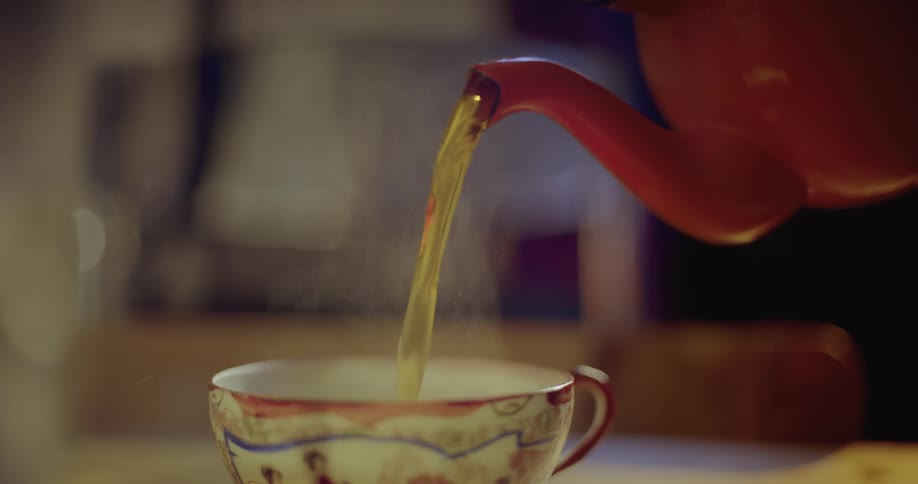 Steaming tea pouring from a orange teapot into a china cup