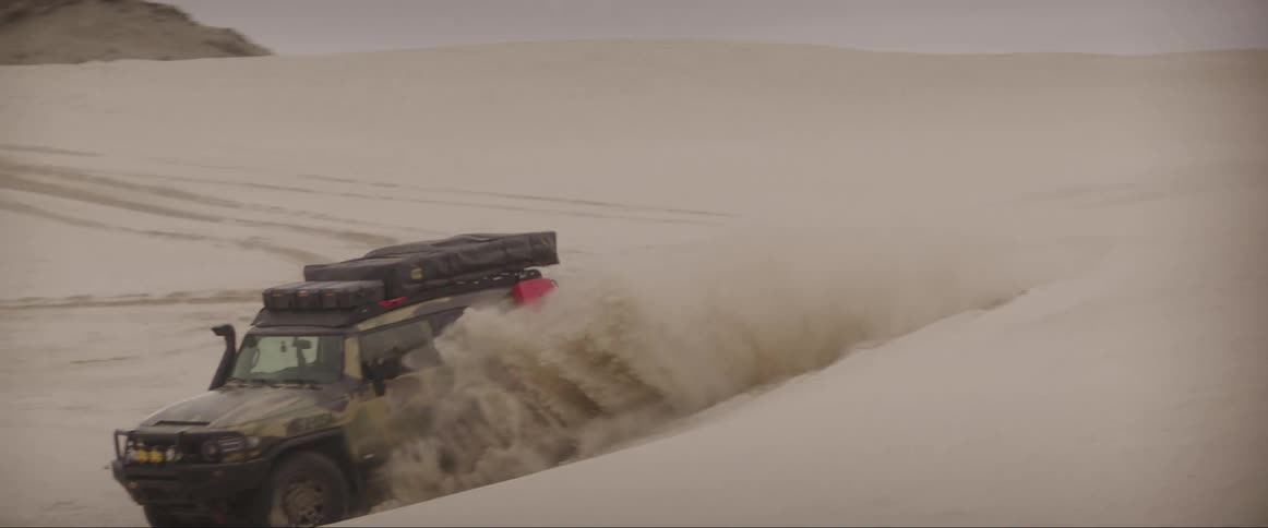 A vehicle drives on sand dunes leaving a trail of sand behind it