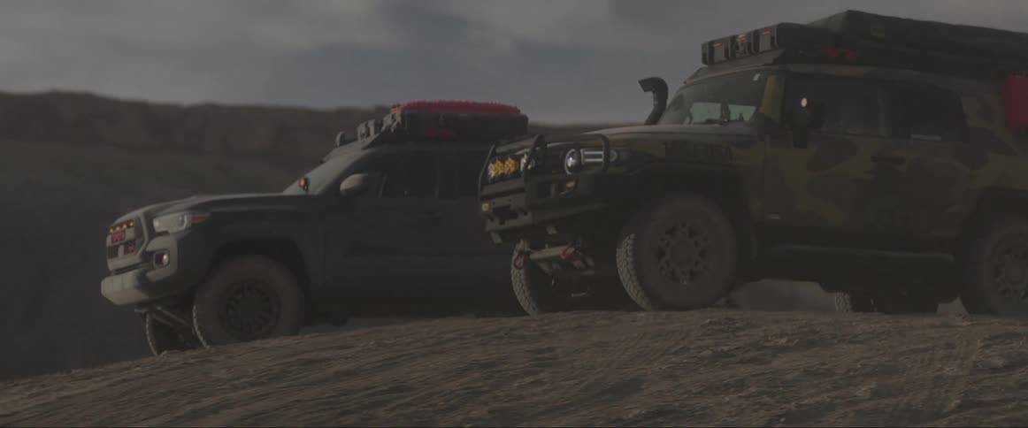 Dirty vehicles sit parked on arid ground near cliffs on a cloudy day