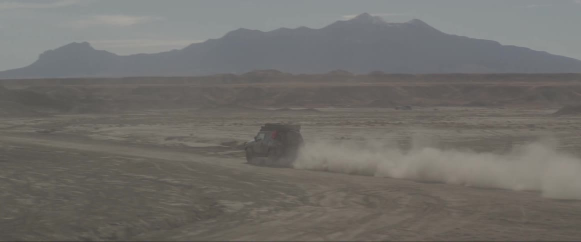 Two vehicles speed along dirt road through desert bordering mountains creating thick trail of dust