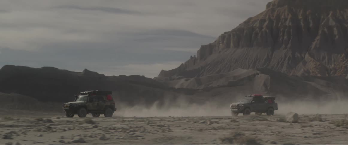 Two offroad vehicles drive on a desert terrain with a mountain in the background