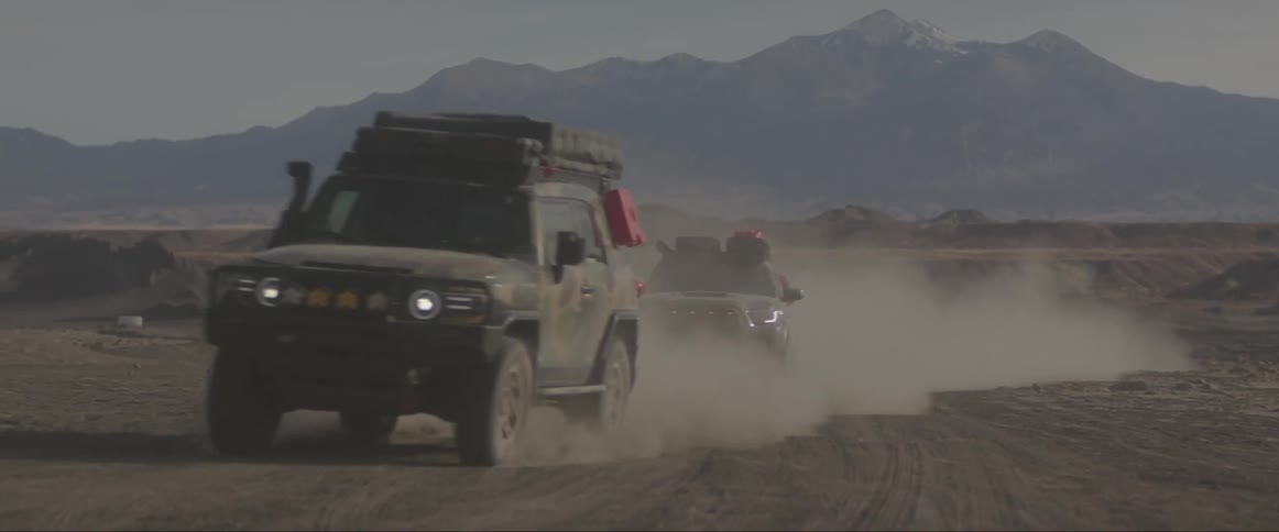 Vehicles drive away from mountain through arid hilly terrain creating a cloud of dust