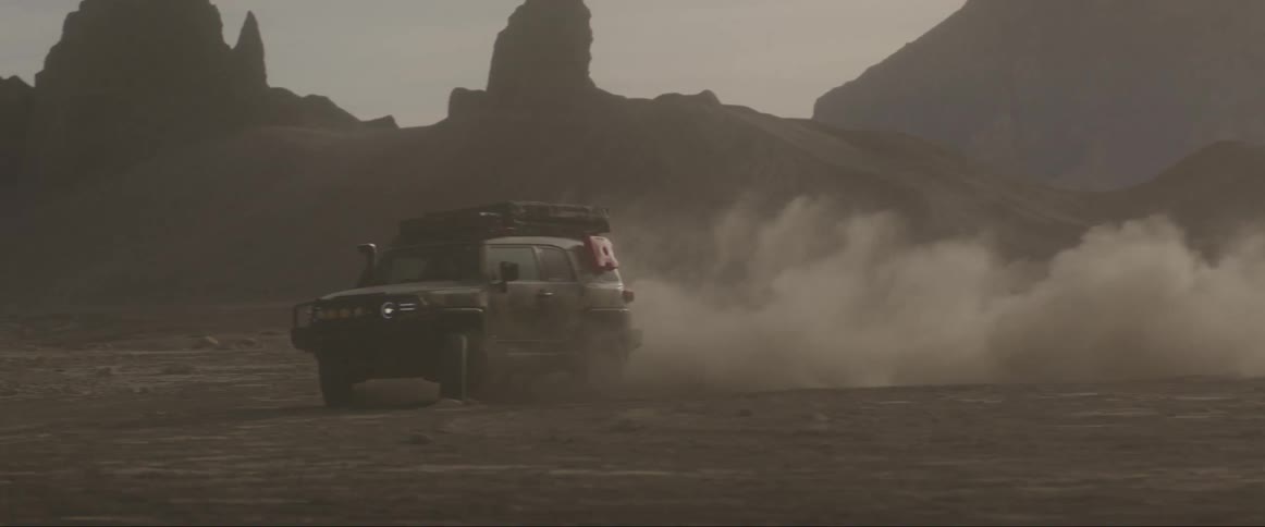 Vehicle drives across desert near badlands creating a cloud of dust