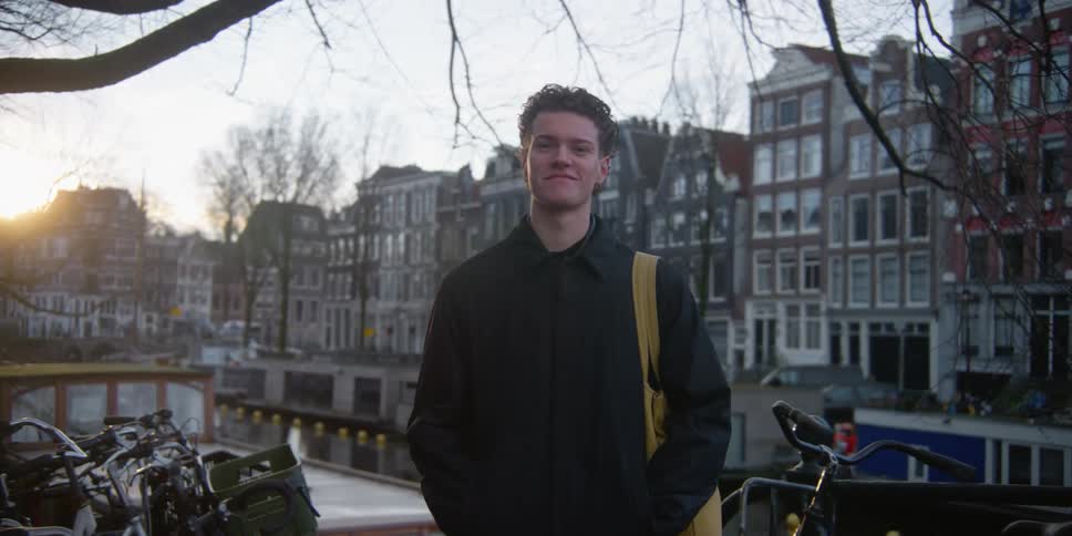 A teenage boy with a bag smirks while standing near bicycles