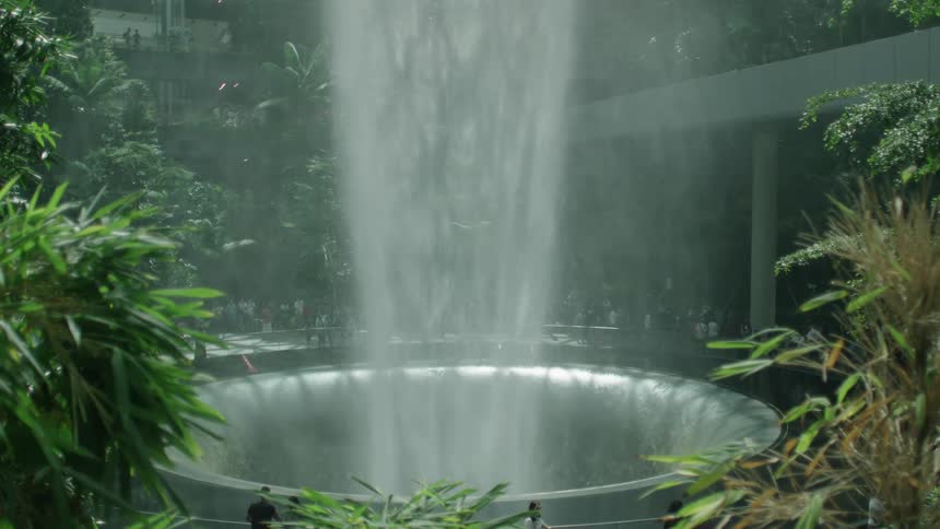 Fountain with water flowing down in the middle of a botanical area with plants and people around