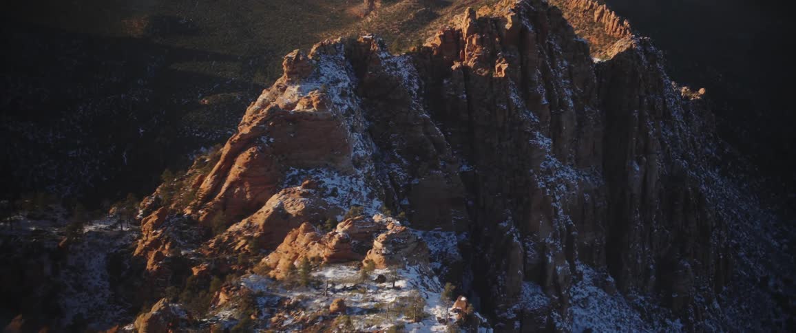 Aerial over a ridge of brown rugged mountain peaks lightly dusted with snow at sunset
