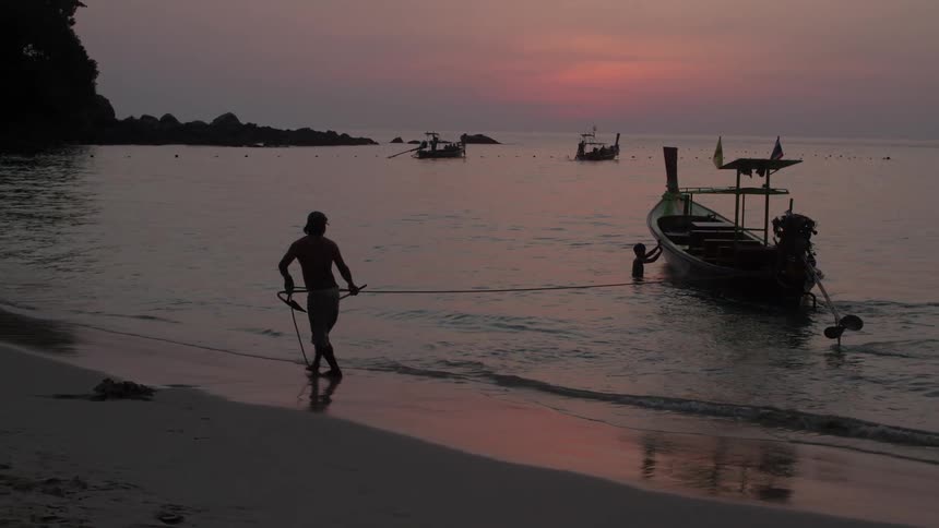 A man on a Thailand beach at dusk uses a rope to land a long boat