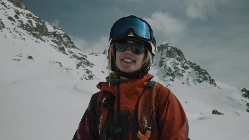 A blonde woman with a helmet and warm orange clothes is smiling and behind her is a mountain covered in snow