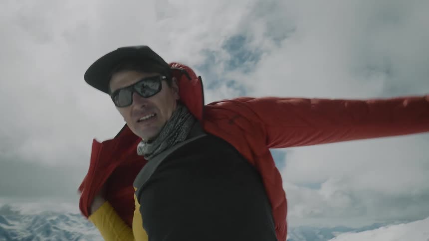 A man standing atop a snowy mountain puts on a winter jacket
