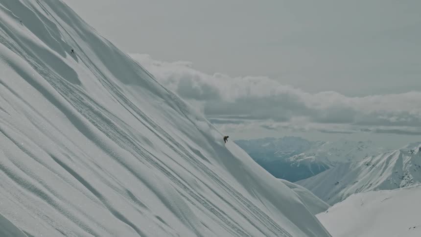 Skier riding a steep backcountry slope down a mountain during the day with mountain peaks and clouds in the background