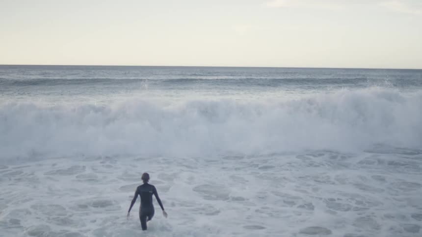 A person wearing a black wetsuit enters ocean with white waves and clear sky