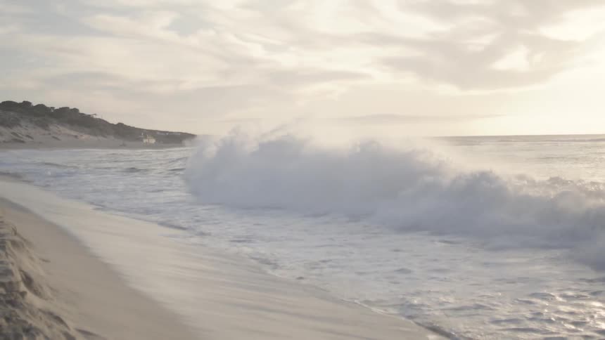 A wave collapses and crashes upon a sandy beach under a sunny sky