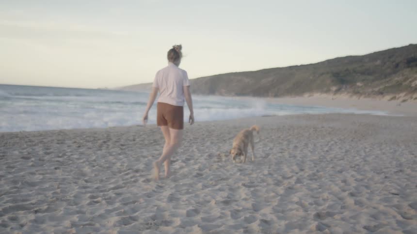 Young adult female walking on beach and throwing a ball to a golden retriever puppy on an overcast cloudy day with a coastal hillside in the background