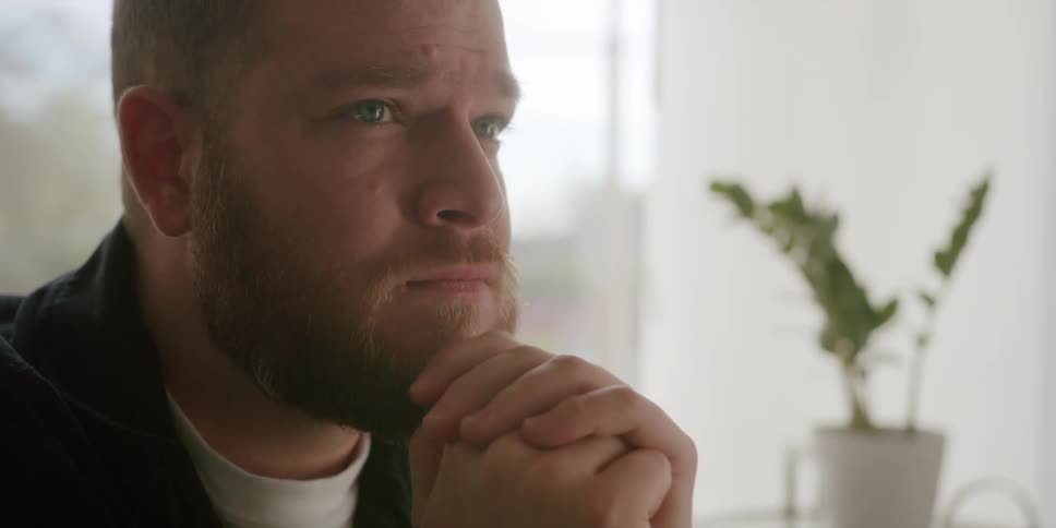 A man sitting in a living room clasps his hands and thinks