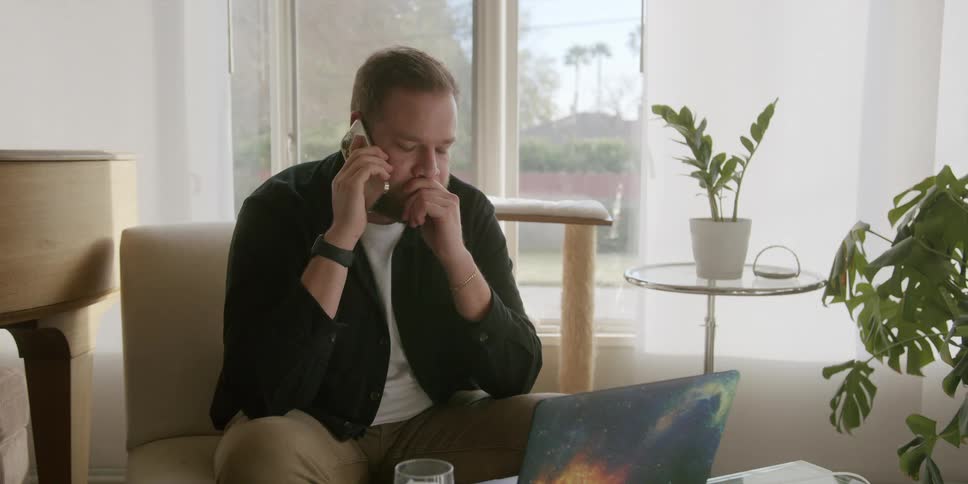 A man is talking on his phone and looking at his laptop