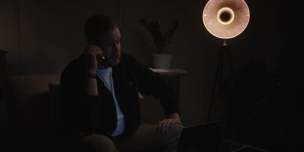 A man sitting alone in a room lit only by one dim talks on his smart phone while looking at a laptop getting progressively more frustrated