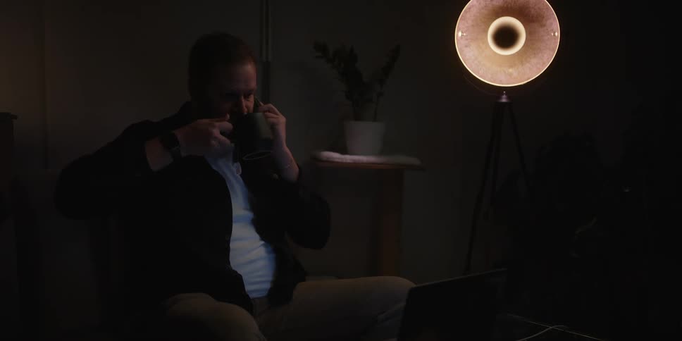 A man sitting alone in a room lit only by one dim lamp drinks tea and talks on his smartphone as he looks at a laptop