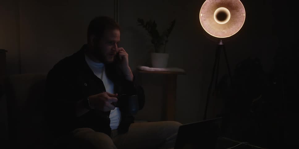 Man sitting in a dimly lit room talking on the phone and holding a cup of tea