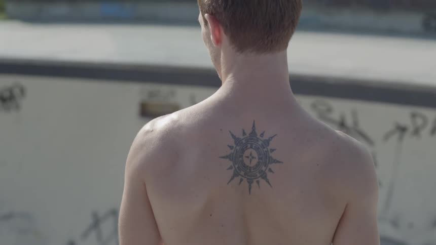 A man with a tattoo on his back stands in front of an empty pool with graffiti