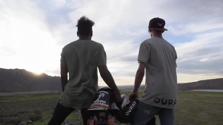 Two skateboarders stare off into an open field holding their skateboarding equipment