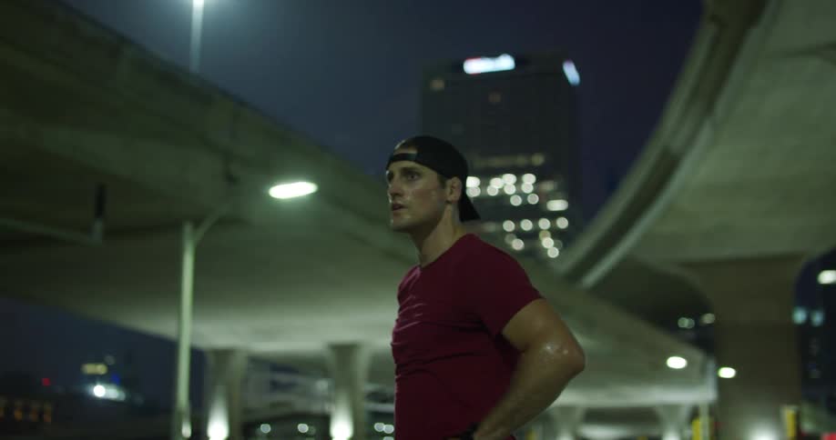 Man with his hands on his waist below an overpass in a city at night with his body sweaty